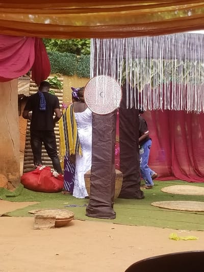 The scene, set in the city of Ochadam, Nigeria, depicts an outdoor or semi-outdoor decorated event space. Overhead, translucent brown fabric forms a canopy, with a section of red fabric draped on the left. To the right, numerous white vertical strings hang from a dark overhead structure. The background features green artificial foliage and a light-colored slatted wooden screen.
Three individuals are present. On the left, a male figure, seen from behind, wears a black t-shirt and black pants, with a blue and white patterned cloth draped over his left shoulder. He stands near a light brown structure. In the center, a female figure, also viewed from behind, wears a white lace dress and a purple and yellow striped headwrap/shawl, positioned near the male. To the right, another male figure, facing away, wears a dark shirt and blue jeans, walking towards the right side of the frame.
The ground consists of light brown earth in the foreground, transitioning to a green artificial grass mat. On the green mat, a large red fabric-wrapped object is visible near the left male figure. Several round, flat woven mats are spread across the green matting. Two dark, fabric-wrapped cylindrical pillars stand vertically, one adorned with a circular woven decoration displaying a spiral pattern. A small, round woven basket rests on a terracotta-colored stand in the left foreground.