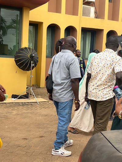 The scene is an outdoor paved area in Madina, Ghana, situated in front of a warm yellow-orange two-story building. The building features rectangular windows, some reflecting palm trees, and decorative arched recesses on the upper floor, with an air conditioning unit visible. The ground is covered in reddish-brown interlocking pavers.
In the center, a bald, dark-skinned male is viewed from behind, walking, wearing a light gray short-sleeved button-up shirt, light blue jeans with horizontal seam details, and white and burgundy Reebok sneakers. He carries a black shoulder bag on his left side and wears a beaded bracelet on his right wrist. To his left, a black parabolic softbox with yellow ribs is set up on a silver tripod stand, alongside a black equipment bag on the ground. Another similar softbox is partially visible behind him.
To his right, partially obscured figures include a person in a light-colored long garment with patterned slides, and a dark-skinned male in a white patterned shirt and khaki pants, holding a plastic water bottle. In the mid-ground, a dark-skinned male with a beard is visible, wearing a dark suit with a blue and yellow patterned ceremonial stole, observing the area. The front of a dark grey vehicle is partially visible in the bottom right.