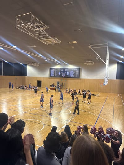 A medium-shot image taken at an indoor basketball court in Lausanne, Switzerland, shows two basketball teams lined up on the court during a game or practice. A scoreboard displays team names, “Les Roches” and “EHI Eagles,” and scores “56” and “38”. Spectators are seated along the sidelines, some with pom-poms. The environment includes overhead lighting, basketball hoops, and standard court markings.