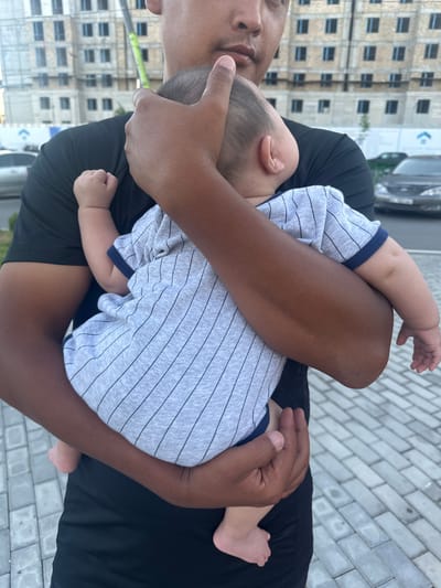 A man holds a baby wrapped in his arms. The baby is wearing a gray onesie with thin black stripes and a blue trim. The man is wearing a black t-shirt. The background shows a building under construction and a street with cars. The ground is paved with gray interlocking bricks. The scene appears to be an outdoor location in the city of Manas, Kyrgyzstan.