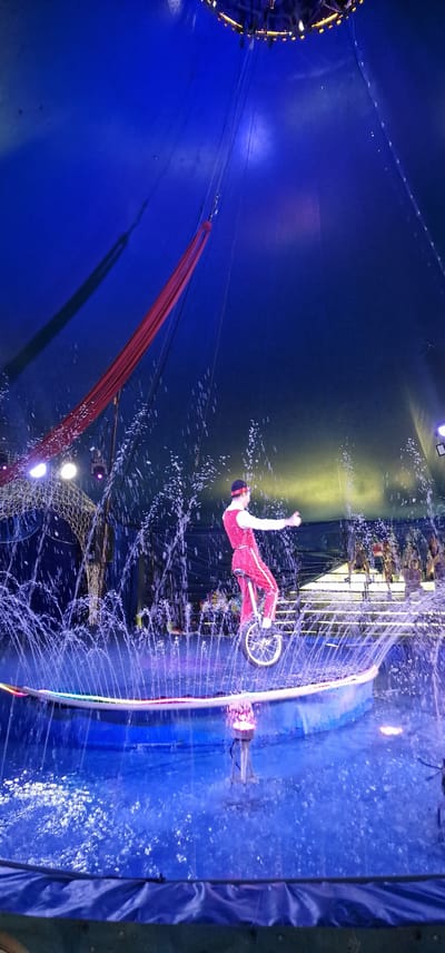 The image depicts a circus performance inside a large tent. A performer in a red costume is riding a unicycle on a circular platform surrounded by water and fountains. The tent ceiling is visible, illuminated by blue and purple lights. Red fabric is hanging from the ceiling, possibly part of the aerial performance setup. The image appears to be taken in Saulkrasti, Latvia, during a circus event.