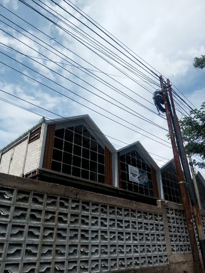 A building with a unique roofline featuring multiple gable-shaped sections sits behind a textured concrete block wall. The building has large, multi-paned windows. An electrical pole and many power lines cross the sky above, with scattered clouds. The color palette leans towards muted grays, browns, and whites, reflecting a typical urban or suburban setting. Part of a tree is visible on the right side.