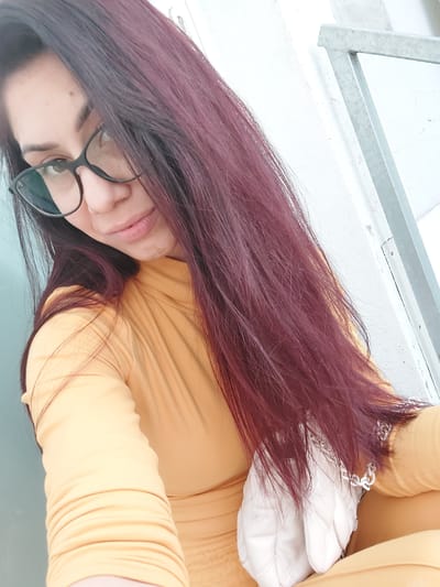 A close-up, high-angle selfie shows a woman with long, dark reddish-brown hair and glasses. She is wearing a yellow turtleneck top and holding a white quilted purse with a silver chain strap. The background is a textured white wall with a light grey metal railing on the right side. The woman is looking down and to the side with a slight smile. The image does not provide enough information to determine the specific location in Altdorf, Germany.