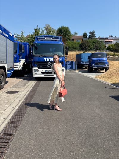 A young woman stands on an asphalt surface in front of a row of blue trucks on a sunny day. The woman is wearing a light-colored, floral-patterned dress and silver heeled sandals. She is holding a small, coral-colored handbag in her right hand and a white cloth in her left. Behind her, the trucks are parked in a lot. The truck on the far left is a silver and blue emergency vehicle with "THW Hilfswerk" written on its side. To its right is a larger blue truck with "THW" emblazoned on the front. Another blue truck, a Mercedes-Benz with a trailer, is parked further to the right. The background shows a dry, grassy embankment, trees, and houses with solar panels on their roofs. The scene appears to be an outdoor area associated with the THW (Technisches Hilfswerk) in Obernburg am Main, Germany, given the visible signage on the vehicles. The weather is clear and sunny with a bright blue sky.