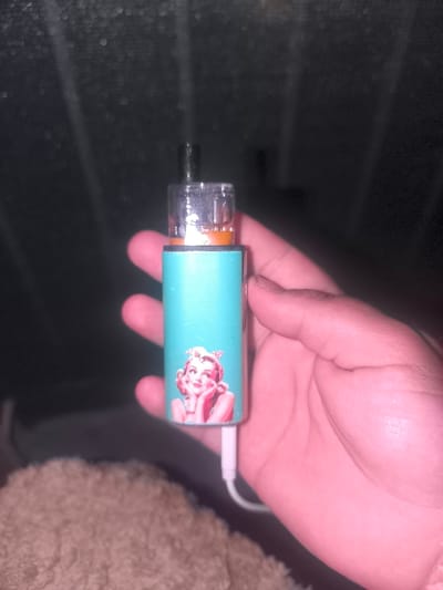 A hand holds a turquoise vape pen with a pin-up girl sticker on it. The vape pen has a clear cartridge with orange liquid at the top. A white charging cable is plugged into the bottom of the vape pen. The background is dark and out of focus, with some bokeh lights visible. The image does not provide any information about the location of Lauterbourg, France.