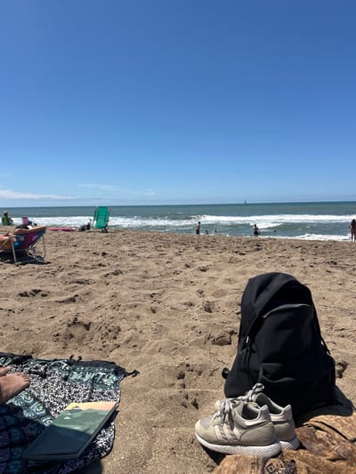 A beach scene with a clear blue sky and ocean. People are in the water and along the shoreline. A black backpack and grey shoes sit on the sand next to a colorful patterned blanket with a laptop on it. Part of someone's feet can be seen near the bottom of the blanket. Other beachgoers are visible in chairs near the water.