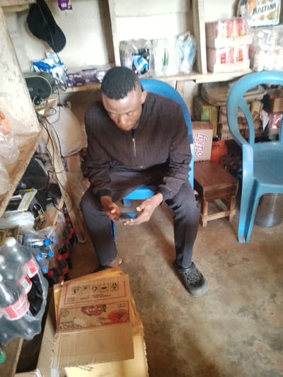 The image shows a man sitting in a room that appears to be a small shop or storage area in Agwala, Nigeria. The man is wearing a dark brown zip-up jacket and dark pants, with dark clogs on his feet. He is looking down at a mobile phone in his hands. The room is cluttered with various items, including shelves stocked with packaged goods such as bottles of soda, boxes of food items, and what appear to be cleaning supplies. There are two blue plastic chairs visible in the scene, one of which the man is sitting on. A small wooden stool is placed next to the man. The environment suggests a functional, likely commercial space where goods are stored and possibly sold. The lighting in the room is somewhat dim, contributing to the overall atmosphere of the scene.
