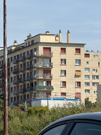 This media file shows a tilted view of a residential building in Antony, France. The building is light beige with many windows, some of which are partially open, revealing red shutters. The architecture appears to be from the mid to late 20th century, with multiple balconies and protruding sections.

In the foreground, a rusty metal railing runs horizontally, suggesting the viewer is observing from a balcony, bridge, or similar elevated structure. To the right of the building, dense green foliage, possibly a large bush or tree, obscures part of the scene. The sky is clear and pale blue, indicating daylight.

The image's orientation is such that the building appears to be leaning or tilted, which is likely due to the camera's angle and the way the photo was captured. There is no discernible activity or event taking place. No people are visible. The lighting suggests it is daytime, with no specific indicators of the time of day other than the general brightness. The location cue is provided by the context of Antony, France. There is no visible text in the image.