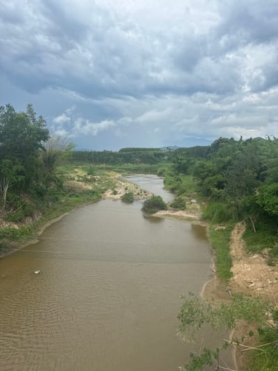 The image is a high-angle shot of a river flowing through a lush, green landscape. The river has muddy brown water and its banks are lined with vegetation and sandy soil. In the foreground, a net is stretched across the river, appearing to be a barrier or fishing net. The river winds through a valley with trees and foliage on both sides. In the distance, rolling hills covered in trees are visible under a dramatic sky filled with dark, heavy clouds, suggesting an approaching storm. The overall impression is one of a natural, somewhat wild environment, likely in a rural or semi-rural area. The location is identified as Suối Câu, Vietnam.