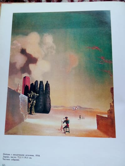The image is a painting depicting a surreal landscape. In the foreground, a figure dressed in a green jacket and red sash sits on a stool, facing a canvas on an easel. The figure wears a black beret. The landscape is vast and desolate, with a sandy ground and distant, hazy mountains. To the left, there is a dilapidated structure with a red tower and a cloaked statue standing in front of a group of dark, pointed trees. In the far distance, several small, white, cloud-like forms are visible. The sky is a gradient of light yellow and greenish-blue, with some reddish streaks. A portion of text below the painting identifies it as "Landscape with Mysterious Details, 1934. Wood, oil; 72.5 x 59.5 cm. Private collection." The painting does not depict any recognizable landmarks or features of the city of Zhmerynka, Ukraine.