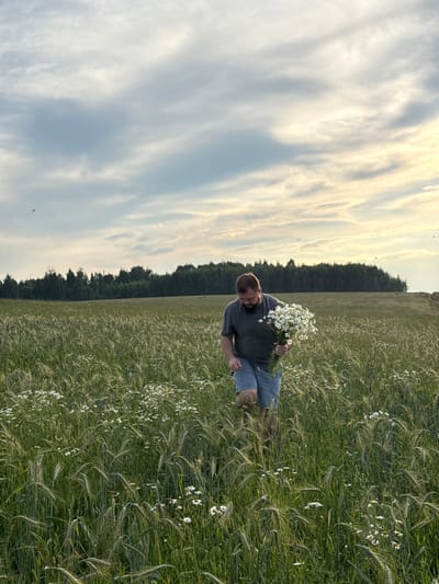 A man is walking through a field of green wheat and wildflowers, carrying a bouquet of white daisies. He is wearing a dark grey t-shirt and blue denim shorts. The field is bordered by a dense line of trees in the background. The sky above is cloudy with patches of sunlight. The scene appears to be located in a rural area, possibly in or near the village of Сновицы, Russia.