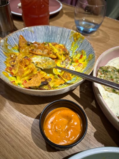 The image displays an indoor dining table scene. In the foreground, a black, ribbed ramekin holds a vibrant orange-red dipping sauce. To its immediate right, a portion of a light-colored bowl is visible. Dominating the midground is a partially consumed dish of nachos in a ceramic bowl with a blue and white striped rim. The nachos consist of tortilla chips coated in melted yellow cheese, black beans, diced red peppers, and green herbs. A silver spoon rests in the nacho mixture, extending towards the right. To the right of the nacho bowl, a pink-rimmed bowl contains a white, creamy dish topped with green herbs and red flakes. Behind this, a clear glass with a blue-tinted base is present. In the background to the left, a red beverage in a textured glass and a silver condiment shaker are visible, alongside a blurred pink plate. The entire scene is set on a light brown wooden table exhibiting a linear grain pattern. No specific architectural details or signage are visible to determine the exact location within Kalyan-Dombivli, India, beyond being an indoor restaurant or cafe setting. No individuals are fully visible, though human presence is implied by the dining arrangement.