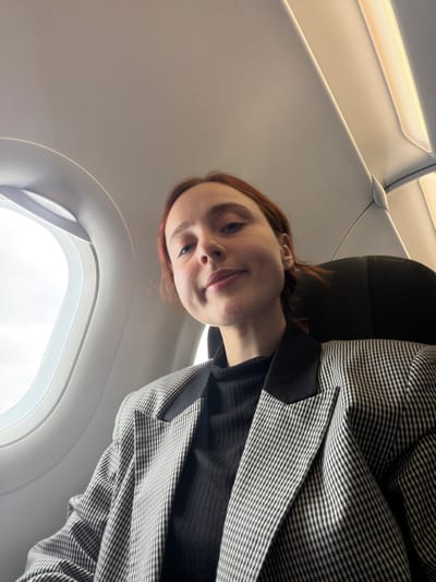 The image depicts a female individual inside an aircraft cabin, consistent with a flight associated with Tuzla, Turkey. She has reddish-brown hair and is wearing a black ribbed turtleneck under a grey-and-white patterned blazer with black lapels. The individual is positioned centrally, looking directly at the camera with a slight smile, exhibiting visible dimples. To her left, an oval airplane window reveals a bright, undifferentiated sky, indicative of daylight. The aircraft's interior features light-colored overhead panels and integrated strip lighting. A dark-colored seat headrest is partially visible behind the individual.