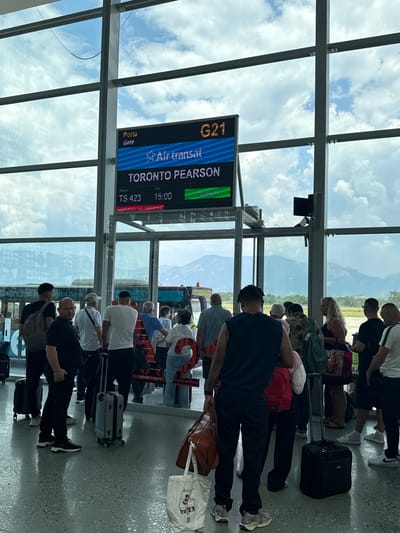 The image depicts a group of people in what appears to be an airport terminal, possibly a departure gate, indicated by the flight information display. The subjects are standing and waiting, some with luggage, suggesting they are travelers. The flight board shows "TORONTO PEARSON" as the destination, with flight number "TS 423" and a departure time of "15:00". The text "Application not registered!" is also visible on the display. The setting features large windows looking out onto a cloudy sky and mountains, with the sky appearing bright, suggesting it is daytime. The interior of the terminal is modern with a tiled floor and metal structural elements. The people are dressed in casual clothing, and their postures do not convey any strong emotions, primarily appearing patient or perhaps a bit weary from waiting. The overall atmosphere is one of transit and anticipation. The mention of "Kruja, Albania" in the prompt provides a specific geographical context, suggesting this airport terminal is located in or near Kruja.