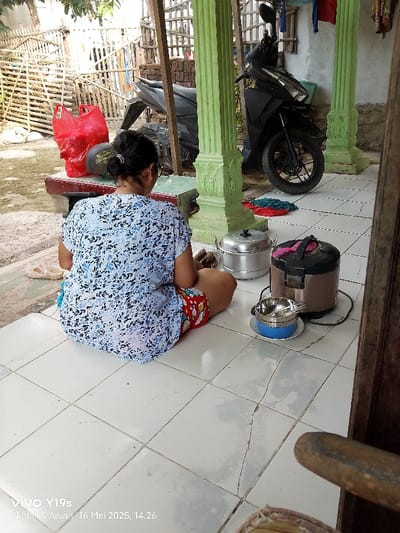A woman is sitting on a tiled floor wearing a blue and white patterned shirt, red shorts, and sandals. Her legs are crossed. A rice cooker and a large pot are visible on the floor near her. A blue bowl with spoons is on the floor next to her. In the background there is a motorcycle. The environment is a house in the city of Gegesik, Indonesia (latitude: -6.538, longitude: 108.43).