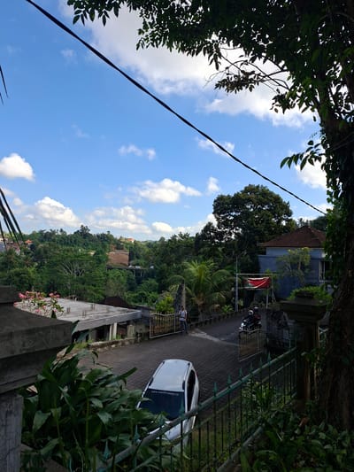 The image depicts a street scene in Banjarangkan, Indonesia, characterized by lush vegetation and a bright, cloudy sky. In the foreground, a gray stone pillar and a dark green metal fence frame the view. Partially visible is the rear of a white car parked on a paved road. Further down the road, a person stands near a gate, and two individuals ride a motorcycle. Buildings with tiled roofs are scattered among dense trees and hills in the background, with a bridge visible in the distance. Overhanging tree branches and a dark power line cross the top of the frame.