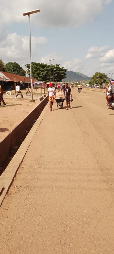 This image depicts a street scene in Gboko, Nigeria, likely during daylight hours under a partly cloudy sky. The road is unpaved and dusty, with a concrete drainage ditch running along the left side. On the right side of the road, two individuals are walking, one pushing a wheelbarrow laden with what appear to be goods, possibly food items, covered in plastic. The person pushing the wheelbarrow wears a white t-shirt and a colorful patterned skirt, with a red headscarf. The other individual, walking beside them, wears a black t-shirt and shorts, carrying a black bag.
In the background, several structures with corrugated iron roofs line the left side of the street, suggesting a market or commercial area. A prominent sign reads "REXHOOD ART S". Further down the road, more buildings are visible, and several people are walking. A few motorbikes are also present, with one carrying two passengers on the right side of the frame. Distant hills are visible on the horizon, suggesting a slightly hilly landscape. The overall atmosphere appears to be that of a busy day in a semi-urban or rural African setting. The presence of a solar-powered street light near the foreground indicates some modern infrastructure alongside traditional elements.