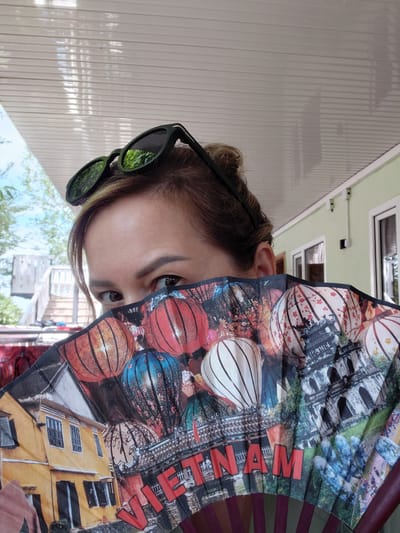 A woman is holding a large, decorative fan in front of her face, with only her eyes and forehead visible. She is wearing dark sunglasses on top of her head. The fan is adorned with images of hot air balloons, traditional Vietnamese architecture, and the word "VIETNAM" in large red letters. The background shows a light-colored, corrugated ceiling, a pale green wall with white trim, and windows. Trees and a wooden structure are visible in the distance. The scene appears to be outdoors, possibly on a porch or patio, in a location that suggests a warm climate. The city of Ақши, Kazakhstan is not depicted in this image.