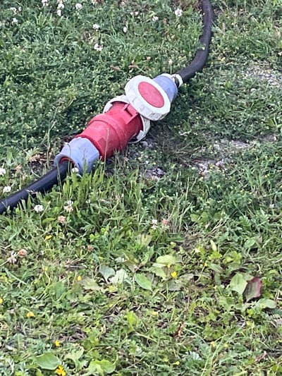 This media file depicts a close-up, top-down view of a red and blue industrial electrical connector lying on the ground amidst green grass and small white and yellow wildflowers. The connector is made of plastic, with the red portion being larger and cylindrical, and the blue portion being a ring around a smaller white circular component. A thick black cable extends from one end of the connector, disappearing into the lower right corner of the frame. The grass is dense and uneven, suggesting an outdoor setting, possibly a garden, park, or rural area. The lighting indicates it is daytime, and the overall scene appears casual and unposed. There is no visible text in the image. The location context provided is Warffum, Netherlands.