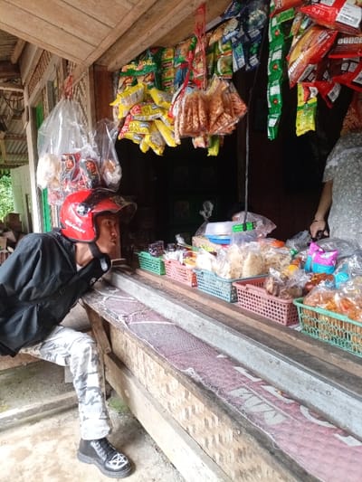 The image shows a young man wearing a black jacket, white pants, and a red motorcycle helmet. He is standing near a wooden structure. The structure appears to be a shop, as it has shelves lined with packaged goods and an open doorway. The goods are mostly snacks, such as crackers and chips. The shop is located in Cisewu, Indonesia, at the coordinates -7.41 latitude and 107.542 longitude. The man is looking at something inside the shop, which is not visible in the image.  The environment is rural and the surface of the shop floor appears to be made of concrete.