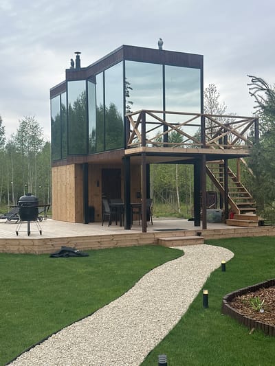 The scene captures a modern glass and wood structure, potentially a house or pavilion, set in a green, outdoor environment. The structure features a two-story design with large glass windows reflecting the surrounding trees and sky. A wooden framework extends above the glass section, hinting at an incomplete or decorative element.
A wooden deck surrounds the structure, furnished with outdoor seating and a grill. A light-colored gravel path winds through the manicured lawn next to the deck. The environment consists of a grassy lawn with garden lighting and a border of mulch.
The image's origin is Vaibla, Estonia. The photograph is taken in the daytime under overcast skies. There are no people visible in the frame.
