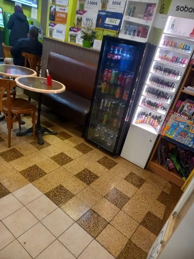 The image shows the interior of a shop or café. A refrigerated display case holds drinks, positioned next to shelving filled with various goods. A brown banquette is accompanied by small, round tables and wooden chairs. Two people are seated at one of the tables. The floor is tiled in a square pattern with alternating brown and light tan tiles.