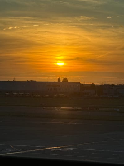 The image captures a sunset over an industrial area, likely near Zaventem, Belgium, given the presence of airport-related infrastructure. The sky is dominated by vibrant orange and yellow hues, with wispy clouds creating a textured pattern. The sun, a bright disc, is visible near the horizon. In the mid-ground, a long, low-rise building with a dark roof stretches across the frame, possibly a warehouse or industrial facility. In front of this building, a line of vehicles, possibly trucks or vans, are parked, their shapes silhouetted against the brighter sky. Further in the background, power lines and sparse trees dot the landscape. The foreground consists of an asphalt surface with painted markings, suggesting an airport tarmac or an industrial lot. The overall scene is one of a developing dusk in an urban industrial environment.