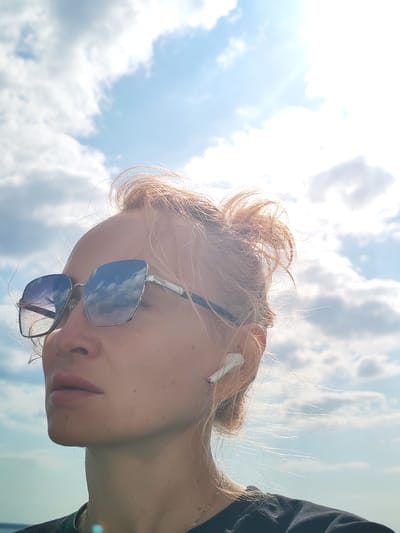 A woman with blonde hair pulled up is shown in profile against a bright, cloudy sky. She is wearing large, square-shaped sunglasses with silver frames, and a white wireless earbud in her right ear. Her hair is light blonde and appears windswept. The reflection of clouds is visible in her sunglasses. She is wearing a dark t-shirt and a silver necklace. The lower part of the image suggests an outdoor setting with a hint of water and land in the distance. The location is identified as Krokhino, Russia.