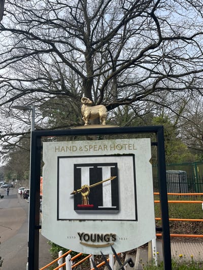 The image captures the entrance sign of the Hand & Spear Hotel, located at Old Dorking Road, Weybridge, Elmbridge, United Kingdom. Central to the frame is a large, light-colored sign with a dark border. The top of the sign reads "HAND & SPEAR HOTEL". Below this, a prominent emblem features a stylized "H" with vertical black and white stripes, overlaid by a golden hand gripping a golden spear. The bottom of the sign displays "YOUNG'S" flanked by "ESTD 1831". Directly above the sign, a golden statue of a ram is positioned.
In the background, a large, bare deciduous tree with extensive branching stands against an overcast sky, suggesting a late winter or early spring season. To the left, a paved road or driveway curves towards the background, where multiple parked vehicles are visible. Several indistinct human figures are observed walking away from the camera on this road. A dark grey streetlight pole extends upwards on the left side, behind the main sign. To the right of the sign, an orange horizontal barrier and green mesh fencing are present. The immediate foreground on the right shows ground cover with patches of grass and small yellow flowering plants. No specific actions or interactions beyond general movement are discernible among the distant figures.