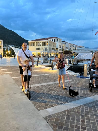 The image depicts an outdoor scene in Kumbor, Montenegro, at dusk. The sky is a deep blue with some clouds, suggesting it is either early evening or late afternoon. The foreground features a cobblestone walkway, and a low wall with a reflective surface.
In the mid-ground, a family is walking along the waterfront. A woman in denim shorts and a black top is holding a leash attached to a small black dog that is walking on the cobblestones. Beside her, a man is pushing a stroller with a young child in it. The child is wearing a hat. Further in the background, there are several buildings with Mediterranean architecture, white facades, and red tiled roofs. Some of these buildings appear to be hotels or apartments. Boats are docked in the water next to the walkway, and there are lights illuminating the scene. In the far background, a dark, tree-covered mountain is visible against the sky.
The overall mood seems relaxed and family-oriented, suggesting a casual evening stroll. The lighting suggests artificial lights are on, indicating it is getting dark.