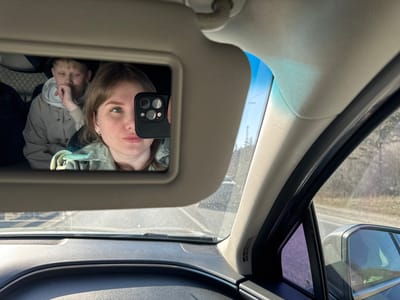 The image is taken inside a car. The subject is reflected in the vanity mirror on the sun visor. The reflection shows a woman holding a smartphone, presumably taking the photograph. Behind her, partially visible in the mirror, is a young boy. The car is moving on a road. The location is Устиново, Russia. The environment outside the car shows trees and a road.