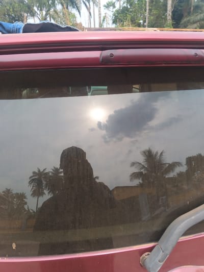 The image depicts an outdoor scene in Itu, Nigeria, viewed through the dusty rear window of a dark red vehicle. The vehicle's dark red body panel forms the bottom and top edges of the frame. A gray rear windshield wiper arm is present in the bottom right corner. Along the top edge of the rear window, a dark red third brake light assembly is visible, fixed to the vehicle's roofline. A folded blue denim-like garment rests partially on the vehicle's roof, visible in the upper-left.
The rear window's surface reflects the immediate surroundings and allows a partial view through it. A prominent, dark silhouette reflection of a person, positioned centrally in the lower half of the window, faces away from the viewer. The reflection also includes a bright sun in the upper-left quadrant of the window, partially obscured by a dark cloud. Other scattered clouds are visible against a lighter sky. Through the window and as reflections, numerous palm trees and other green deciduous foliage are discernible. Reflections of light-colored, possibly single-story buildings are visible in the middle-ground and background. The scene is illuminated by daylight, suggesting a sunny to partly cloudy sky.