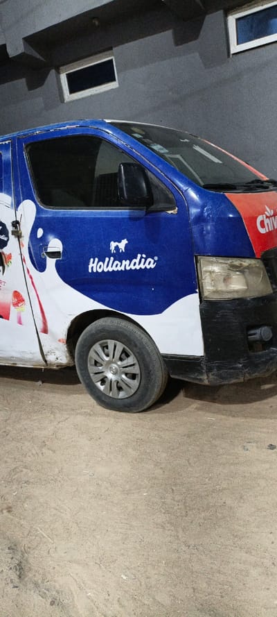 A commercial delivery van is parked on an unpaved, light brown dirt surface in Daurawa, Nigeria. The vehicle's front passenger side is visible, painted predominantly blue and white with "Hollandia" branding and a cow logo on the door, and "Chivita" branding on an orange-red section towards the front. Strawberry graphics and the word "YOGHURT" are visible on a white section further back on the side. The van features a black front bumper, a worn headlight assembly, and a front wheel with a silver-colored hubcap. The bodywork shows signs of wear, including scratches and dirt. In the background, a grey, textured concrete wall with architectural protrusions features two rectangular, dark-paneled openings framed in white. No individuals are present in the scene.