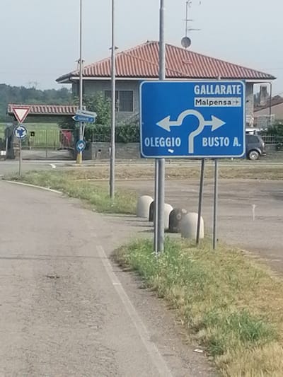 The image shows a street scene with a blue directional sign. The sign indicates that Gallarate and Malpensa are straight ahead, Oleggio is to the left, and Busto A. is to the right. There is a roundabout sign and a yield sign in the background to the left. A house with a red tiled roof is visible behind the directional sign. The scene appears to be an intersection or a point where roads diverge. The environment is somewhat rural, with grass and trees visible. There are no people clearly visible in the image.