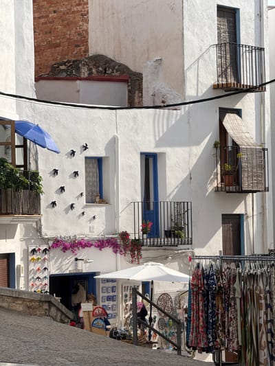 This image captures a vibrant, sun-drenched street scene in Peñíscola, Spain, characterized by classic Mediterranean architecture. The perspective is vertical, looking up a narrow alleyway or street lined with white-washed buildings.
Strong sunlight casts distinct shadows from the architectural elements, indicating a clear, bright day, likely late morning or early afternoon. The buildings feature multiple windows, some with decorative dark metal grates and bamboo-like blinds. Blue frames around windows and a door add pops of color against the white walls.
On the right side, an outdoor shop or market stall is prominently displayed. Racks overflow with folded textiles, including scarves and clothing, in diverse colorful and patterned designs. Below these, a variety of artisanal goods are showcased, such as circular metal wall hangings (including a "Tree of Life" motif), small ceramic pieces, and decorative fans. A large white umbrella provides shade for the merchandise. A striking purple bougainvillea vine cascades down the wall beside the shop's entrance, below which a sign reads "49 ALMA". Further down, a display of small, colorful swallow figures is visible near the blue door.
On the opposing white wall, a unique art installation of numerous black swallow-shaped figures is arranged to appear as if in flight. At the bottom left, a large blue beach umbrella is partially visible, leaning against a railing or wall. A section of rough red brickwork is also exposed on the lower left building facade. Electrical wires traverse the upper part of the image. The scene exudes a lively and charming atmosphere, typical of a historic Spanish coastal town.
