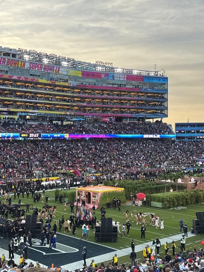 An outdoor stadium is packed with people. On one side, a multi-level structure is decorated with "SUPER BOWL LIX" and other colorful signs. Below, the field is visible with figures performing around a pink structure and hedges. The video screen shows "SAN FRANCISCO BAY AREA" and the time "27:17".