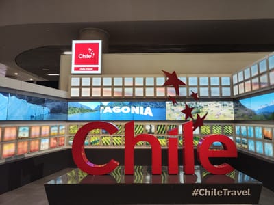 This is an indoor shot of an advertisement for tourism in Chile. It is located inside the city of Pudahuel, Chile. The main subject is a large, three-dimensional red sign spelling out "Chile", along with a hashtag reading "#ChileTravel" underneath. The sign sits atop a glossy black platform, and there are red star-shaped decorations. Behind the sign is a wall of screens displaying landscape photography of Chile, including mountains, lakes, and farmland. Above the screen wall, there is a white sign in red lettering reading "Chile" and "Chile.Travel".