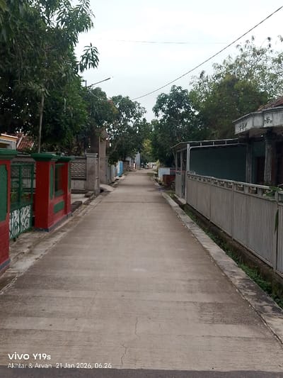 A straight concrete road stretches between houses and fences, disappearing into the distance. Trees line the sides of the road, their branches reaching toward a cloudy sky. A red and green fence stands on the left, while a longer, gray fence runs along the right. The road shows signs of wear with cracks and lines. A small drainage ditch runs beside the gray fence. The bottom of the image contains a watermark.