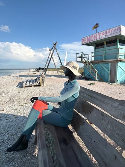 This image depicts a beach scene in Останинское сельское поселение, Ukraine. In the foreground, a mannequin dressed in a teal wetsuit with orange accents and a wide-brimmed hat sits on a weathered wooden bench. The mannequin is positioned to face the left of the frame, with its arms crossed. The beach itself is covered in small white shells.
In the mid-ground, a structure resembling a lifeguard tower or a small building painted teal is visible. It has a sign with pink lettering that appears to say "РЕМОНТ КАЙТОВ" and a phone number. Wooden stairs lead up to this structure. To the left of the tower, a wooden tripod structure stands on the beach, possibly part of a kite or sail rigging. Further back, several vehicles, including what appears to be an SUV, are parked near the water's edge.
The background features a vast expanse of the sea under a bright blue sky with scattered white clouds. The overall impression is of a quiet, possibly seasonal, beach location.