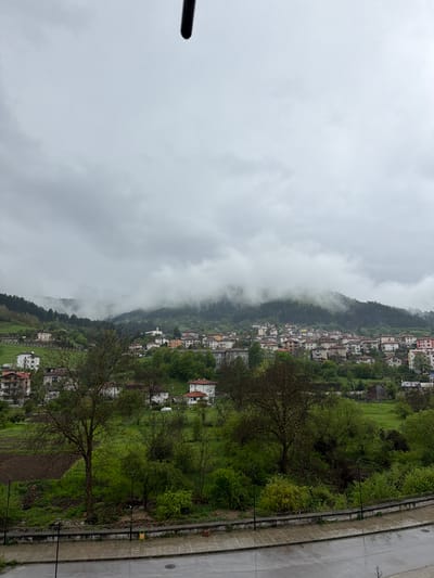 The image depicts a hillside town shrouded in fog, likely situated near or in the city of Devin, Bulgaria. The landscape includes residential buildings clustered along the slope of a hill, interspersed with patches of green vegetation and trees. The sky is overcast with heavy clouds, indicating a damp or rainy environment. Adjacent to the hillside is a paved area, possibly a road or sidewalk, with street lamps. A portion of what seems to be a metal or dark pole is seen at the top left corner.