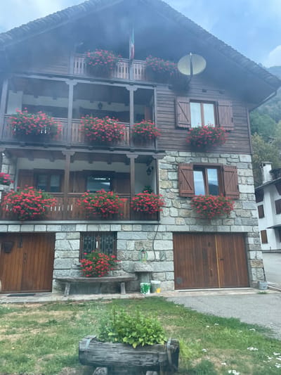 The image shows a multi-story wooden and stone building in Piode, Italy, with a clear blue sky overhead. The building has a dark wooden roof and dark wooden siding on the upper stories, with large stone blocks forming the lower story and parts of the upper stories. There are multiple balconies adorned with abundant red flowers in planters. Several windows are visible, some with wooden shutters. On the ground level, there are two large wooden garage doors and a wooden bench positioned in front of a barred window. In front of the building, there is a grassy area with some white wildflowers and a wooden trough planter in the foreground. The overall scene is picturesque and well-maintained.