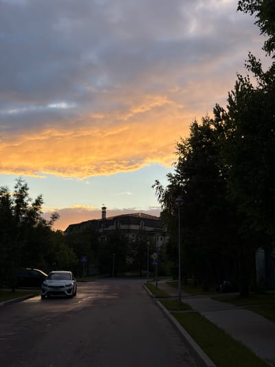 This is a vertical, eye-level, medium shot of a street scene in Zimyonki, Russia, during a sunset. The sky is filled with dramatic clouds, predominantly in shades of grey and illuminated with warm orange and yellow tones from the setting sun.

The scene depicts a road that curves towards the right of the frame. On the left side of the road, there is a sidewalk with a strip of grass and a line of trees. The trees on the far left are lush and dark green, framing the colorful sky. On the right side of the road, there are more trees casting long shadows. Further down the road, a modern white car is parked. The car has a license plate that is partially visible, showing "A218LM". A logo is visible on the car's door, appearing to read "alphagroup".

In the background, on the right side of the road, a building with a clock tower and a domed roof is visible, partially obscured by trees. The architecture suggests it might be a historical or religious structure. There are also streetlights and road signs visible along the street.

The time of day is sunset, indicated by the warm light illuminating the clouds and casting long shadows. The weather appears clear with dramatic cloud formations. The overall mood is serene and picturesque, capturing the beauty of the natural light in a semi-urban setting. There is no visible text in the image other than the license plate and the logo on the car.