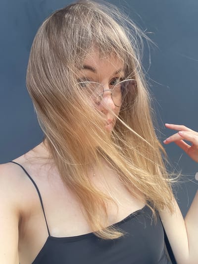 The media file is a portrait of a young woman with long, blonde hair. Her face is partially obscured by her hair, but she is wearing glasses. One of her hands is raised and touching her hair. She is wearing a black top. The background is a dark, solid color, suggesting an indoor setting. The lighting appears to be natural and soft, possibly indicating daytime. There are no visible text elements in the image. The overall mood is casual and relaxed. The provided location context of Zhytomyr, Ukraine, does not offer any specific visual cues within the image itself.