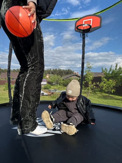 The image depicts a child wearing a beige beanie, dark jacket, and shoes, sitting on a trampoline. Next to the child, a person holds an orange basketball. A basketball hoop with a red backboard is attached to the trampoline net. The setting is outdoors, with trees and buildings visible in the background. The location appears to be Vysokinichi, Russia.