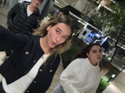 The image captures four individuals in an outdoor urban setting at night in the city of La Purísima, Mexico. In the immediate foreground, a female with light brown wavy hair, wearing a navy blue jacket with gold buttons over a white top and a gold hoop earring, directly faces the camera with puckered lips. To her right and slightly behind, a second female with dark brown hair, dressed in a white turtleneck top, also displays a similar lip posture. In the blurry background, a male wearing a black puffer jacket over a white shirt is partially visible on the left, exhibiting a smile. A fourth individual with dark hair is partially obscured behind the foreground female. The environment consists of modern buildings with illuminated windows, planted trees with sparse leaves, and low-lying greenery on a paved surface. Artificial lighting fixtures are visible throughout the scene. A white vehicle, possibly a utility or commercial vehicle, is partially visible in the mid-background right, adjacent to a dark, slatted architectural element on the far right.