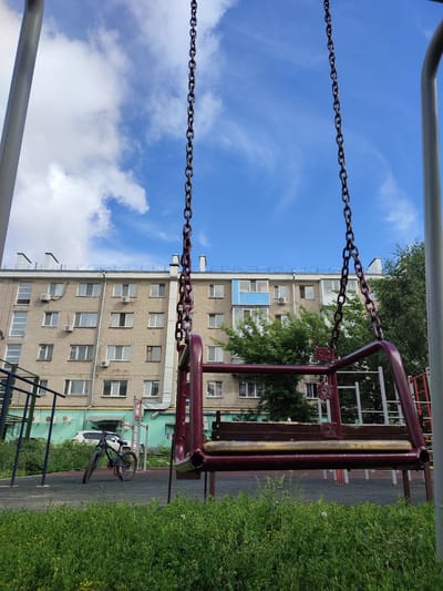 The image depicts a playground scene in an urban setting, likely in Petropavl, Kazakhstan. In the foreground, a red metal swing set is the central object, with its chains hanging down against a bright blue sky dotted with white, wispy clouds. The swing seat itself appears to be made of wood with metal framing.
Behind the swing set, a multi-story apartment building is visible. The building is constructed of light-colored brick and features numerous windows and air conditioning units. To the left of the building, a small green awning covers part of the ground floor. A white car is parked in front of the building, partially obscured by greenery.
To the left of the swing set and in front of the building, a bicycle with blue accents is leaning on the ground. The playground area is surfaced with dark material, and bordered by lush green grass with small yellow flowers in the foreground. Trees are visible behind the apartment building, adding to the urban greenery.
The scene appears to be set during the daytime, with clear, bright sunlight casting shadows. The overall mood is calm and ordinary, suggesting a typical day in a residential area. There is no visible text in the image. The presence of the swing set and apartment buildings strongly suggests a residential courtyard or a small local park.