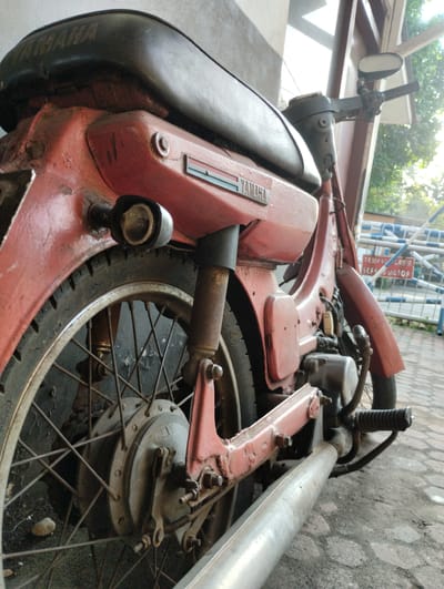 This is a close-up, low-angle shot of the rear of a red, vintage Yamaha motorcycle parked outdoors on a paved surface. The motorcycle shows signs of age and wear, with faded paint, rust, and chipped sections. The focus is on the rear wheel, fender, exhaust pipe, and the lower portion of the seat and body. In the background, a white and blue striped barrier and a sign with red lettering that says "TEMPAT PARKIR SEPEDA MOTOR" (Motorcycle Parking Area) are visible, indicating the location is a designated parking area for motorcycles. The overall lighting suggests it is daytime, possibly late afternoon due to the sunlight filtering through trees in the background. The scene is set in the city of Tanjungsari, Indonesia. There are no people or actions depicted in the image, only the stationary motorcycle and its environment.