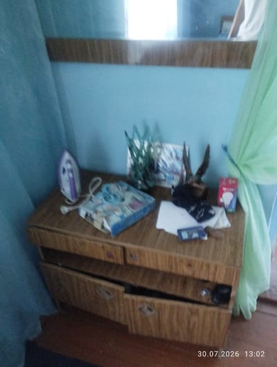 The image depicts an indoor scene, likely a room in a house or apartment, located in Quyosh, Uzbekistan. The primary focus is a wooden dresser with several drawers, some of which are open, revealing contents that are not clearly discernible. On top of the dresser, various objects are placed. These include a purple and white iron, a book or box with colorful illustrations, a mounted animal head with antlers, a small rectangular object that appears to be a phone or remote, a white piece of paper, and a red box containing what looks like a light bulb. To the left, a light blue curtain hangs, and to the right, a light green curtain is draped. The background shows a light blue wall and a reflection in a mirror or window. The date and time stamp "30.07.2026 13:02" is visible in the bottom right corner of the image. There are no people visible in the image.