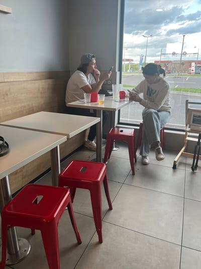 A photograph taken indoors shows two individuals seated at a white table in a brightly lit space. A woman, wearing a light-colored sweatshirt with "NY" written across it, light blue jeans, and white sneakers, is seated on a red stool and leans over the table, looking at her phone. A man, wearing a light-colored shirt and a baseball cap, sits across from her, also looking at his phone. The table holds several clear plastic cups, one with a yellow liquid. Additional red stools are positioned nearby.
Large windows reveal an exterior cityscape with a "Total Energies" gas station visible in the distance, along with a "ONCF" sign. The location is Andrézieux-Bouthéon, France, as indicated by the recognizable gas station and the ONCF sign. The interior has gray tiled flooring, a wooden accent wall, and white walls. A highchair is visible near the window.