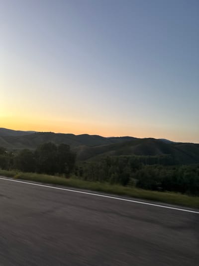 The image depicts a scenic landscape with rolling hills and a vast sky. In the foreground, a paved road with a white line marking the lane stretches into the distance. The road appears to be wet or reflective, suggesting recent rain or damp conditions. The road surface shows motion blur, indicating the photograph was taken from a moving vehicle.

Beyond the road, a strip of green grass and trees lines the roadside. The middle ground is dominated by a series of dark green, undulating hills covered in vegetation. The hills create a layered effect, with some appearing closer and more detailed than others, which fade into the distance. Scattered trees are visible on the slopes of these hills.

The background is a gradient of colors in the sky, transitioning from a pale yellow and orange hue near the horizon to a soft blue at the top of the image. This color gradient suggests the photograph was taken during either sunrise or sunset. The sky is clear, with no visible clouds.

The image does not contain any people, animals, or buildings. The overall environment is natural and undeveloped.

The location is identified as the city of Severnoe, Kazakhstan. However, the visual elements in the image (rolling hills, undeveloped landscape, paved road) do not provide specific landmarks or features that would definitively confirm this location without external information. The scene is characteristic of many rural or semi-rural mountainous or hilly regions.